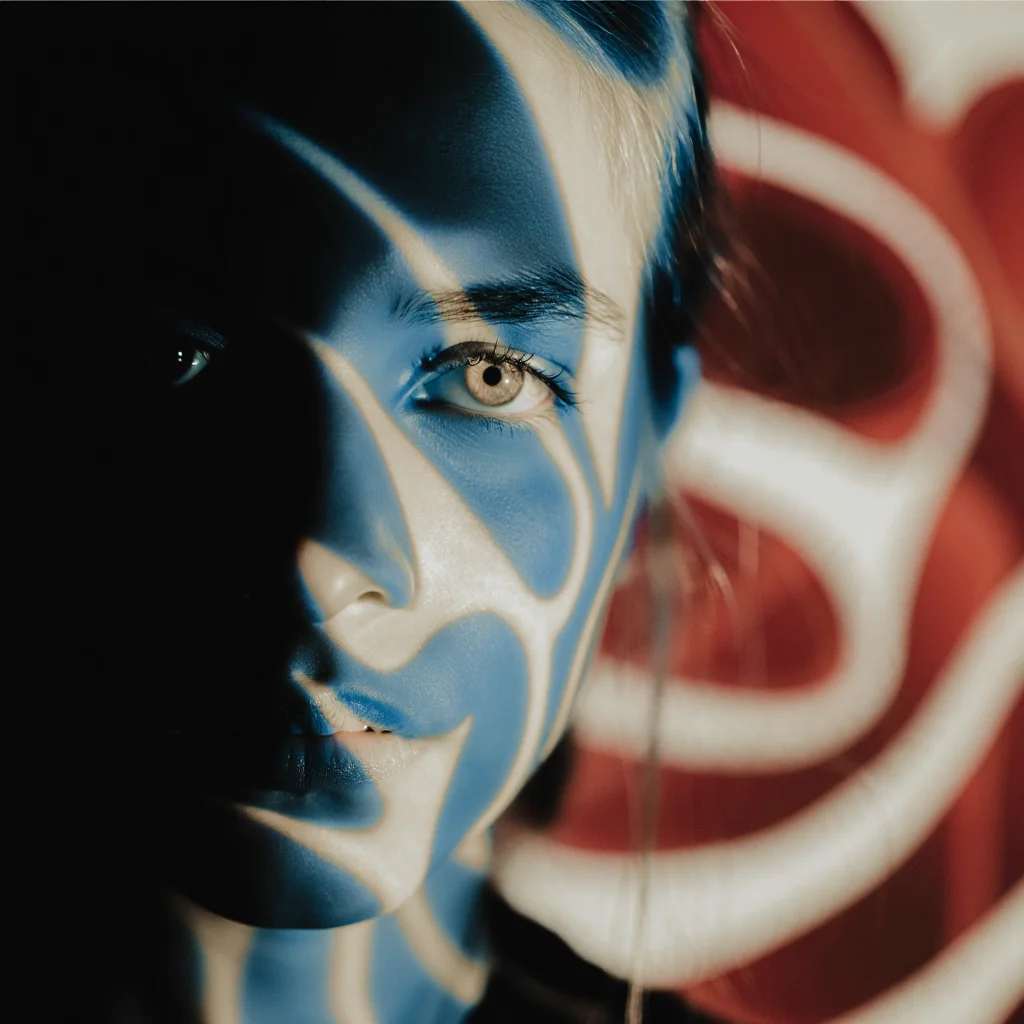 An ethereal blue and sepia toned portrait, centered on the subject's eye peering from the left side of the frame, partially obscured by shadow. The composition uses a shallow depth of field, with the background a swirl of abstract shapes in deep red and white. The overall effect is one of mystery and intrigue, as if the subject is looking out from a dream.